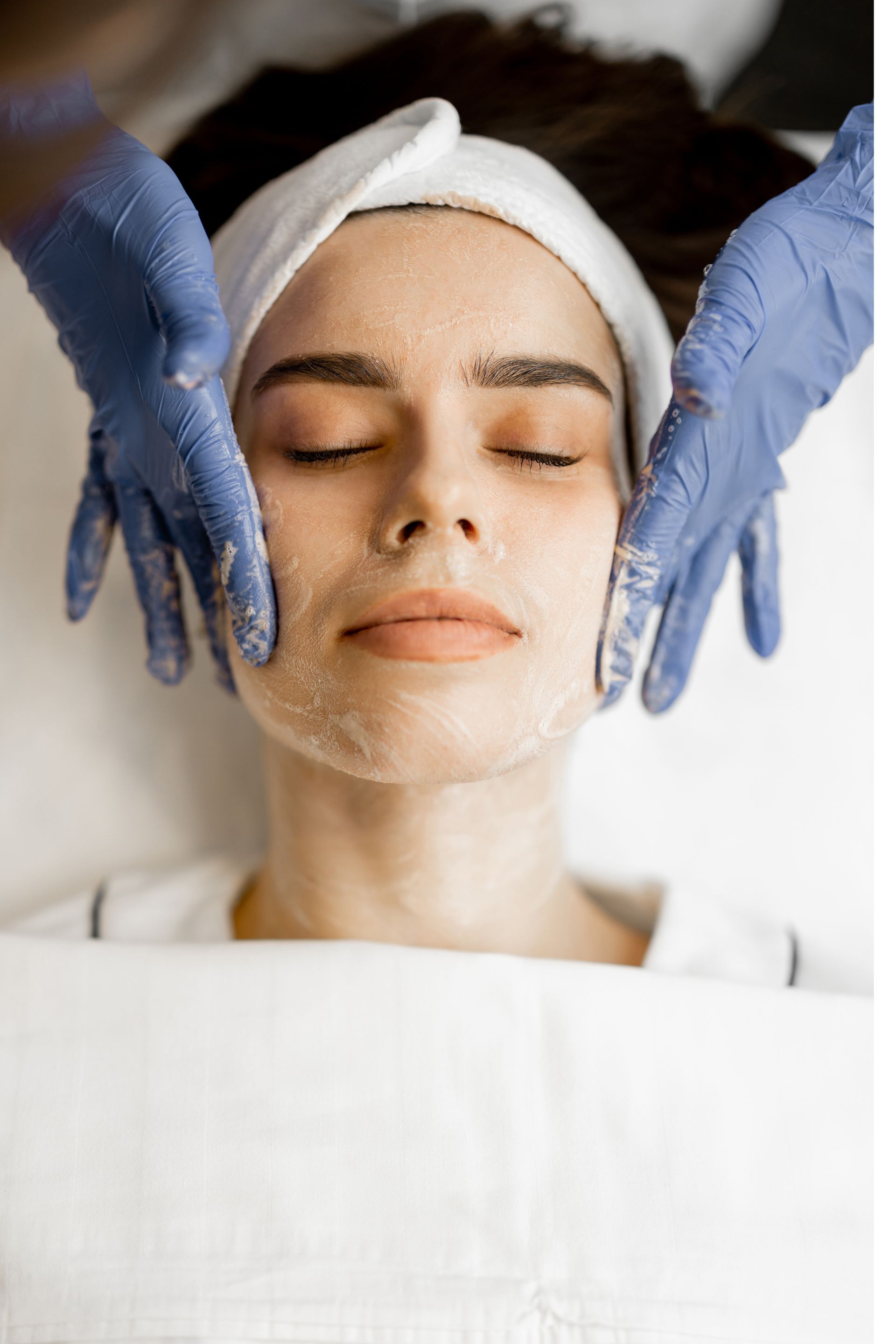 A person with a face mask and headband receives a facial massage from a professional wearing blue medical gloves.