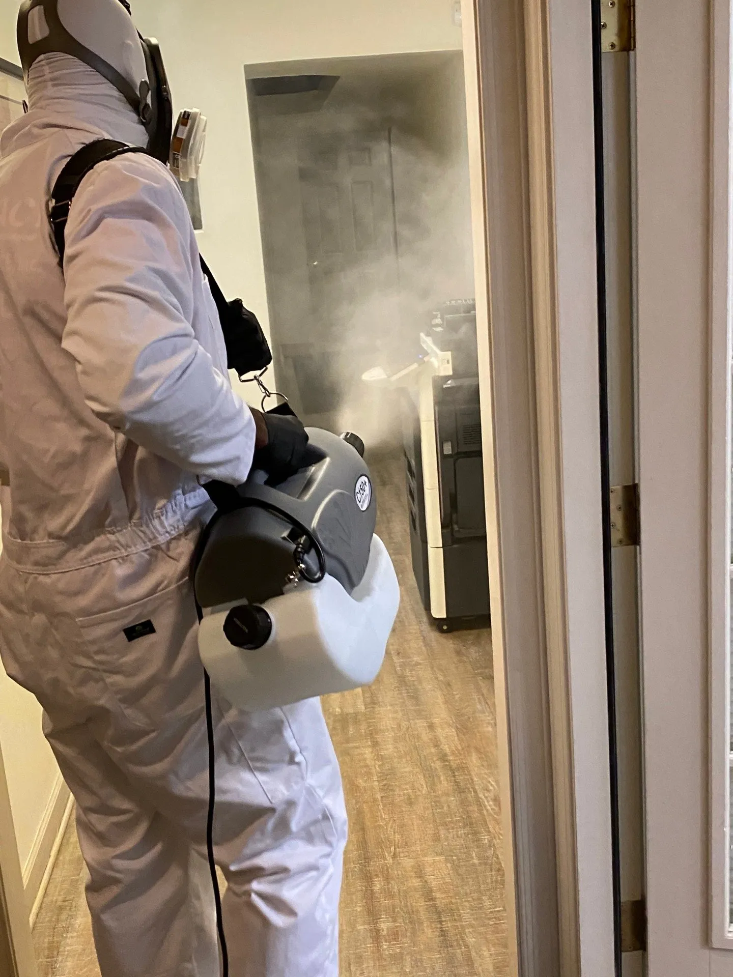 Person in protective suit using fogging machine to disinfect a room.