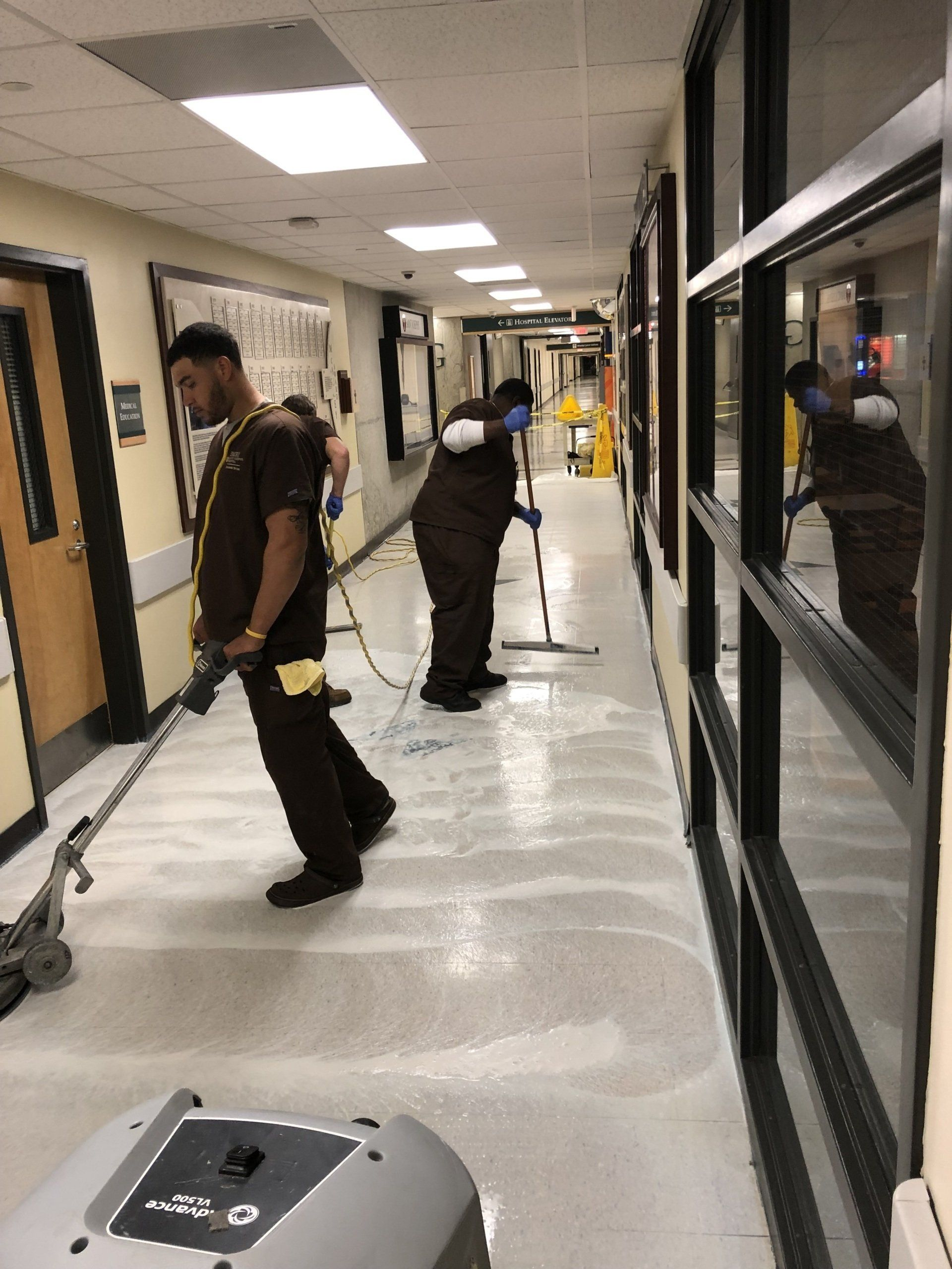 Two people in brown uniforms cleaning a hospital hallway with floor scrubbers.