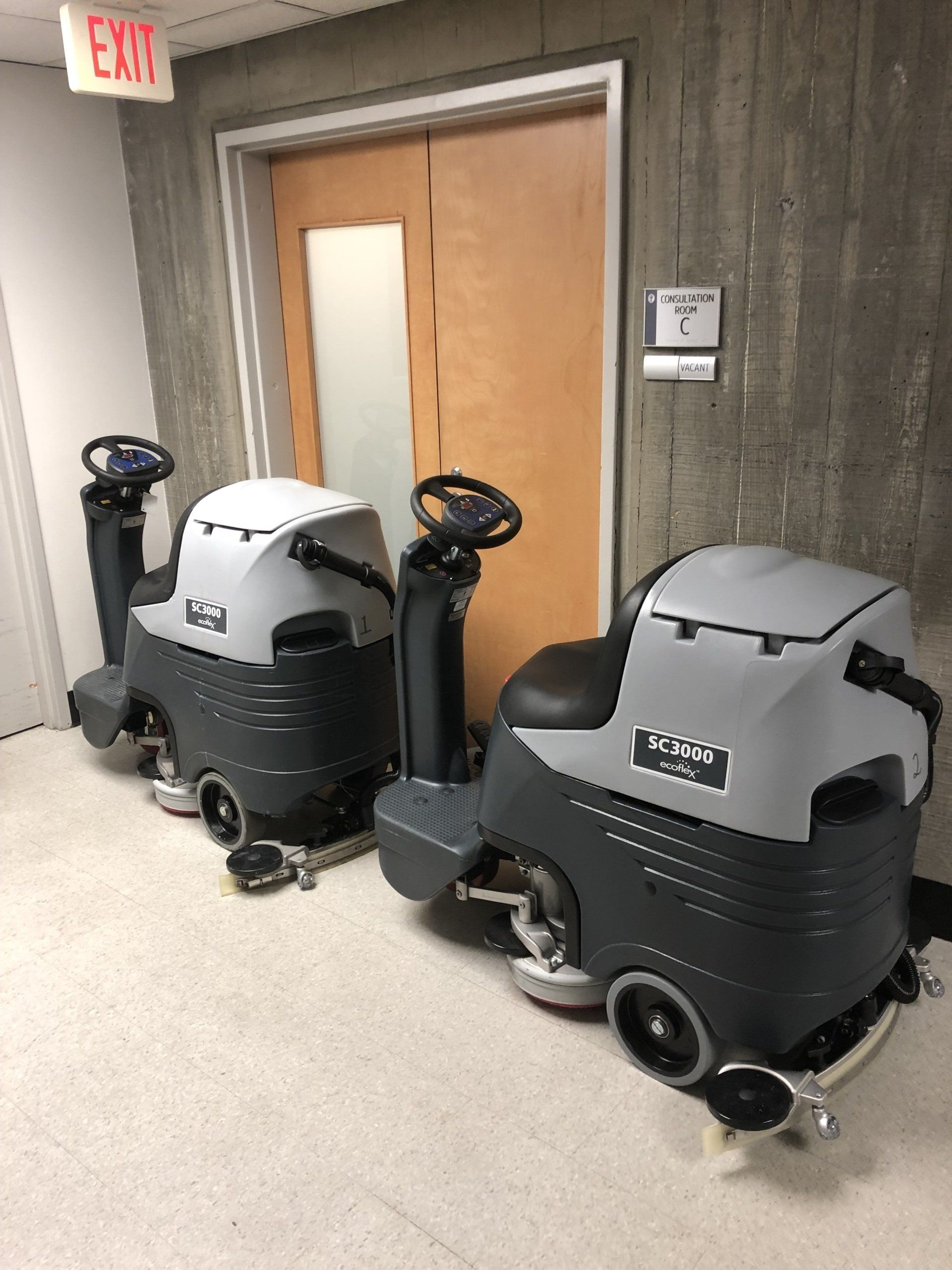 Two gray and black floor scrubbers parked in a hallway with a wooden door and exit sign.