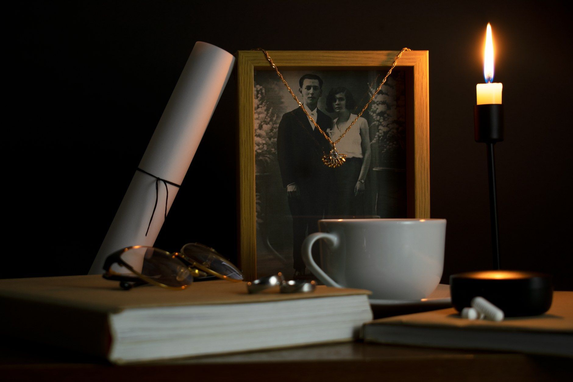 A dark still-life on a wooden table shows a lit candle, a white teacup, a rolled scroll, reading glasses, a stack of books, and an old framed photograph of a couple, all set against a black background.