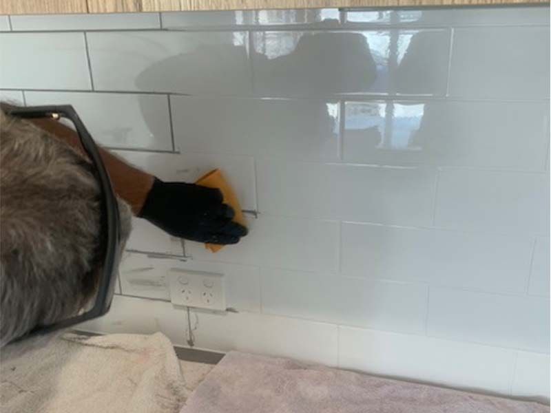 A Man is Cleaning a White Tile Wall With a Sponge — The Tile & Stone Specialists in Peregian Beach, QLD
