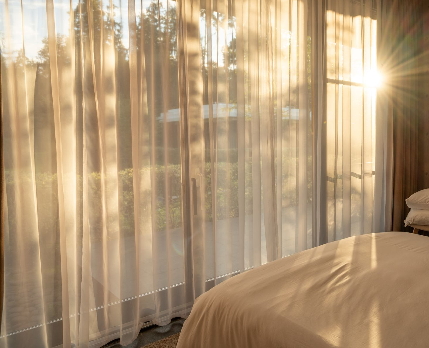 Sunlight streams through sheer curtains in a bedroom, illuminating a bed — GV Blinds in Shepparton, VIC
