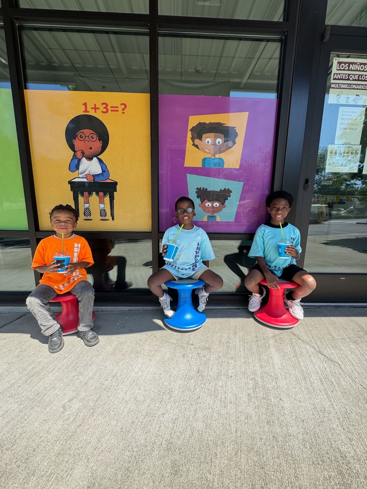 Three young boys sit on stools outside, smiling. Behind them are colorful math posters.