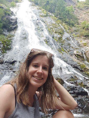 Woman smiling in front of a waterfall, wearing sunglasses on her head, gray tank top, with hand in hair.