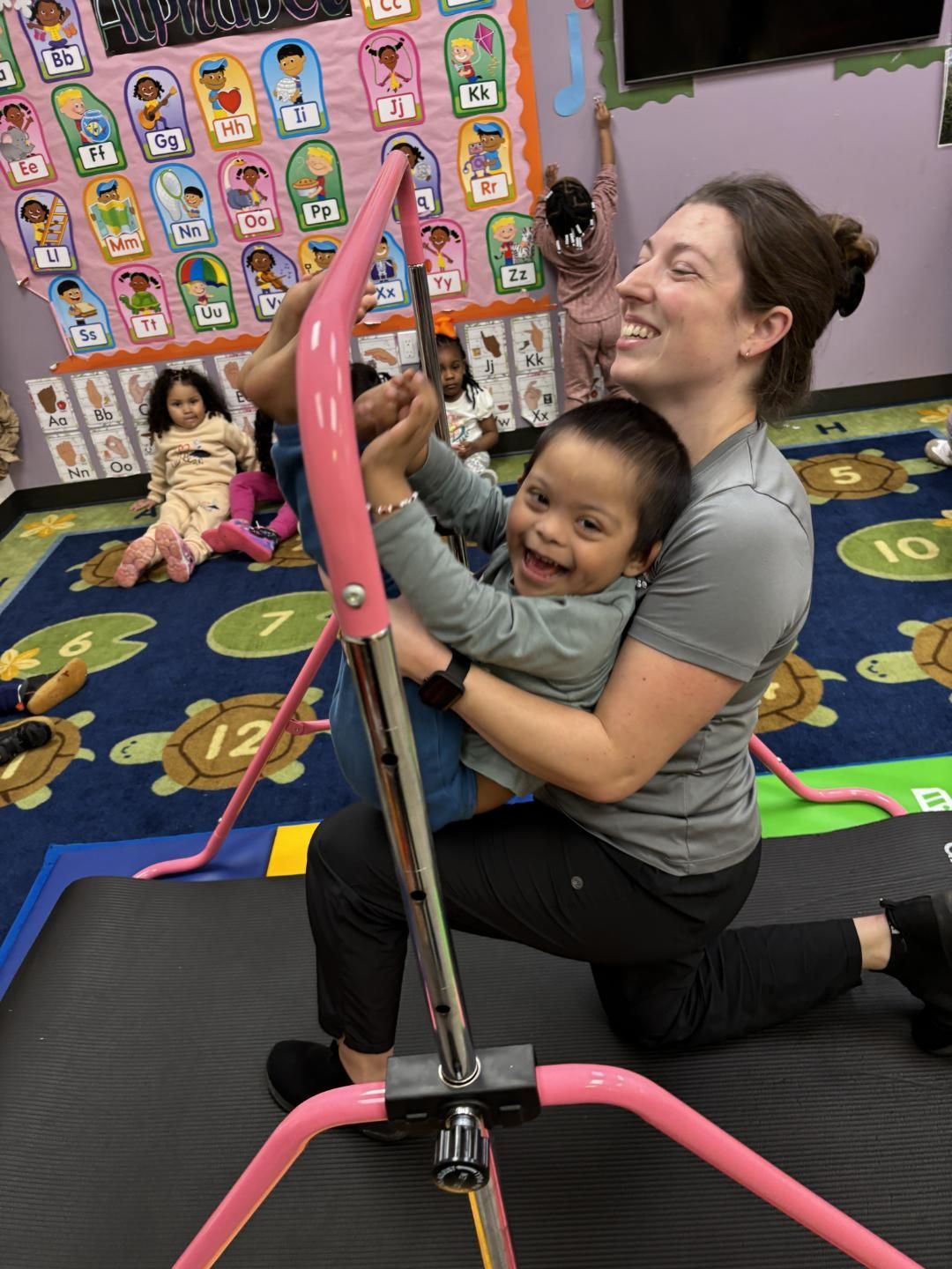 Woman helping a smiling child use pink support equipment in a colorful classroom setting.