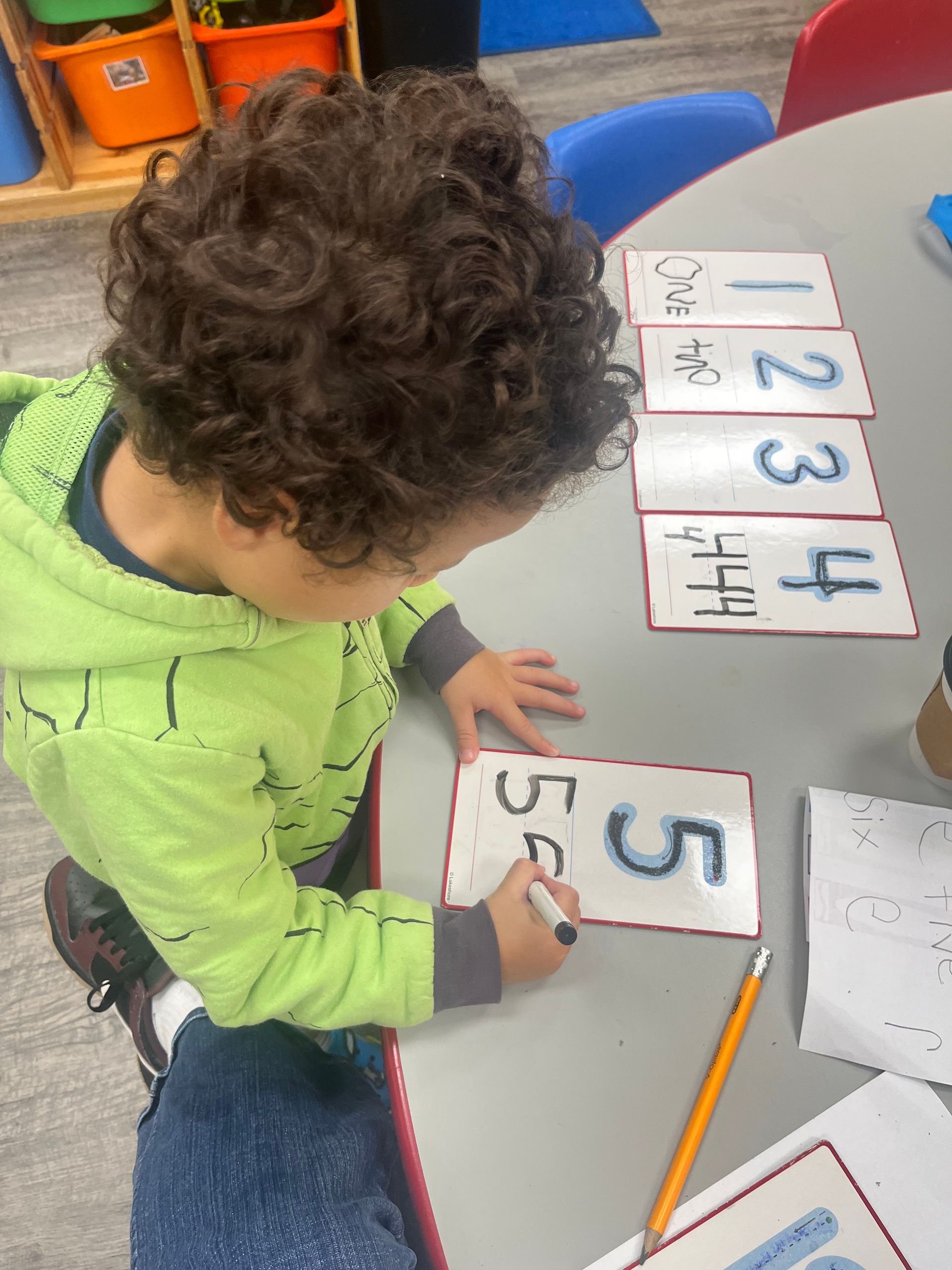 Young child writing the number five on a card, other number cards on a table.