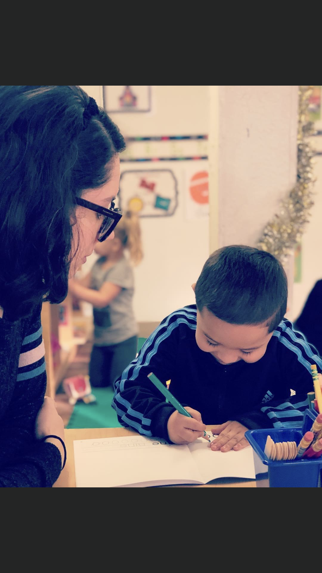 Teacher helps a young boy write at a desk; other children and decorations in the background.