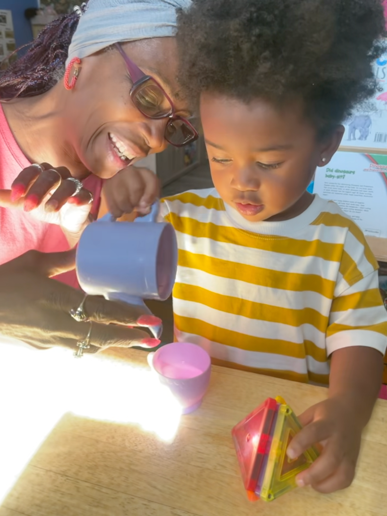 Woman pouring liquid into cup for child, both smiling. Yellow-striped shirt, toys on table.