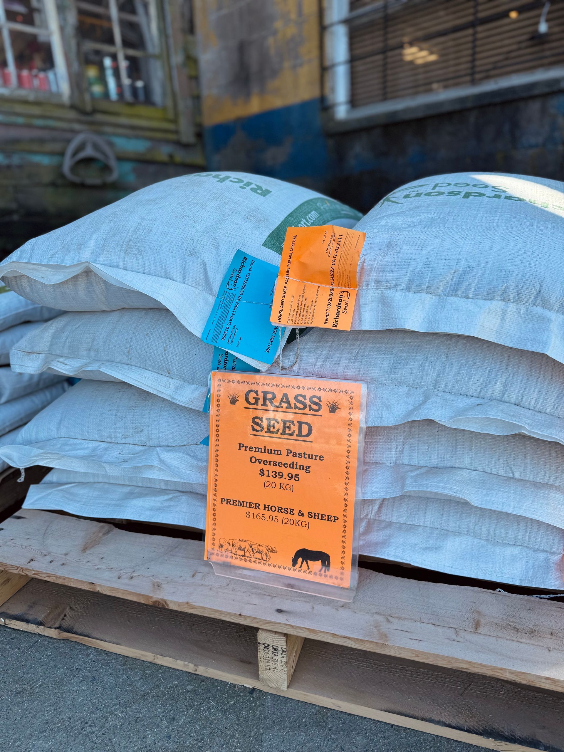 Stacked white sacks of grass seed on a pallet, labeled with orange and blue tags in a warehouse.
