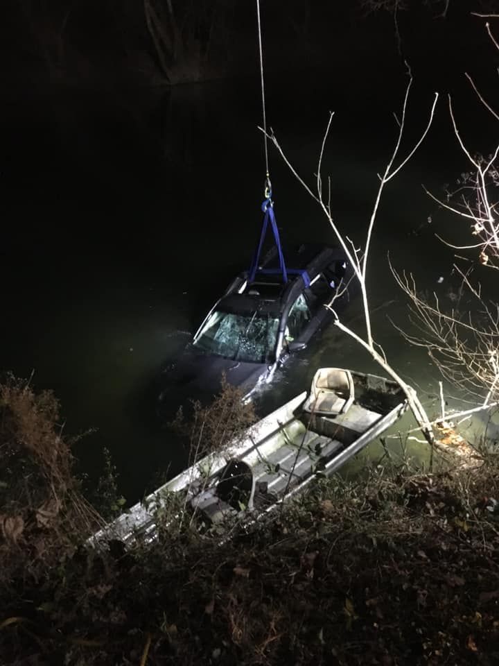 A car submerged in dark water, being lifted by a crane. An aluminum boat sits nearby, at night.