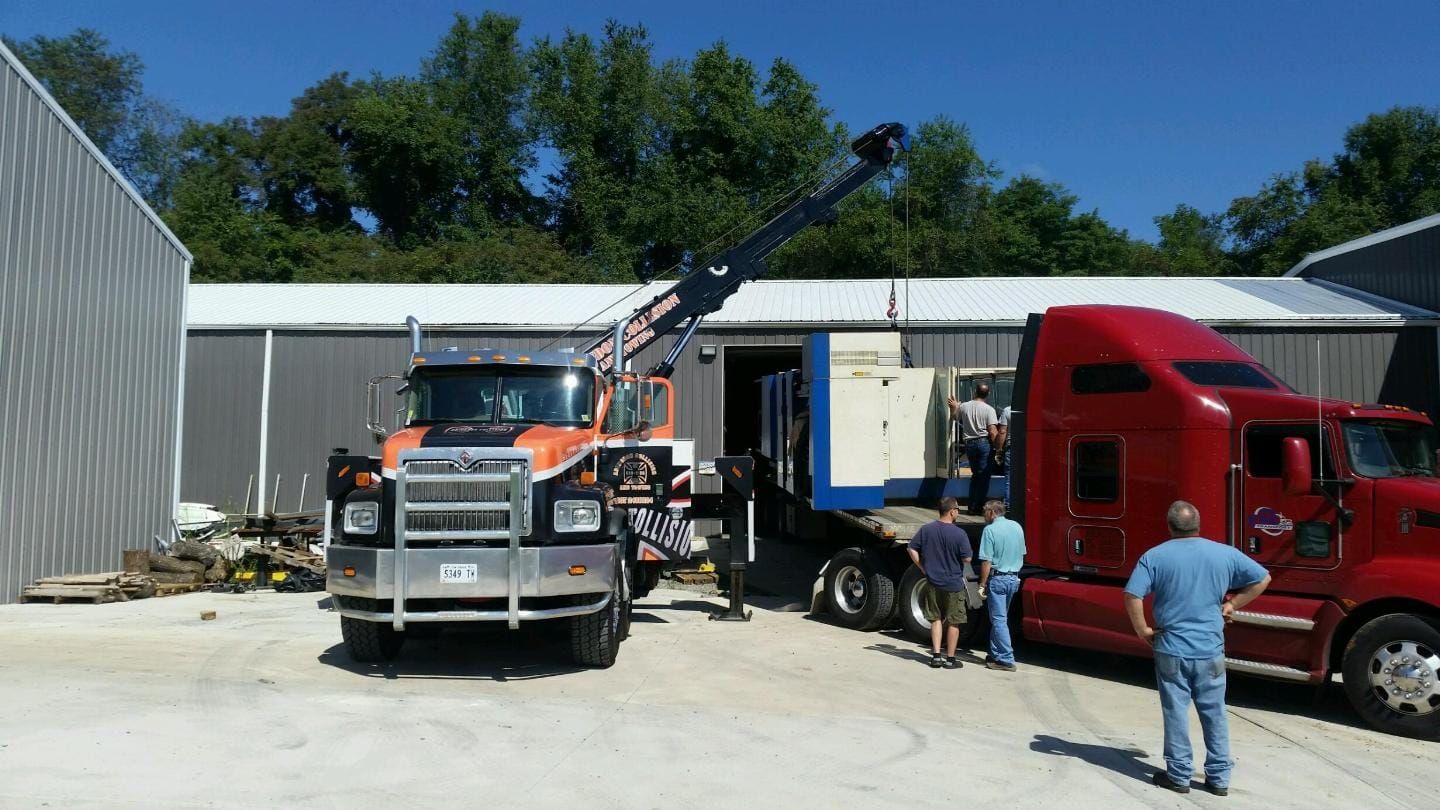 Orange crane truck lifting equipment from a flatbed semi-truck in front of a gray building.