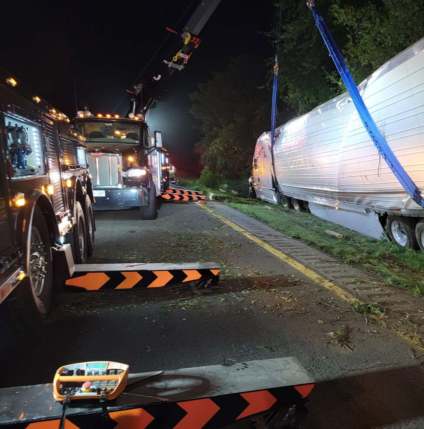 Tow truck recovering a semi-trailer from roadside at night, using a crane and straps.