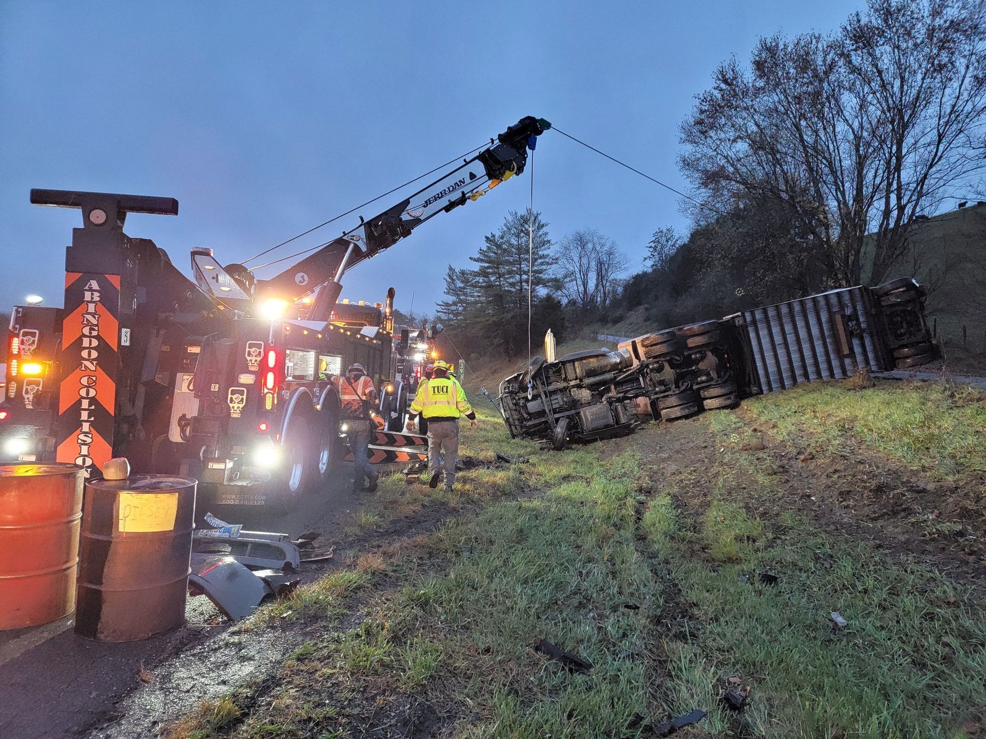 Tow truck recovering a semi-trailer that rolled off the road. The truck is lit up, a worker stands nearby.
