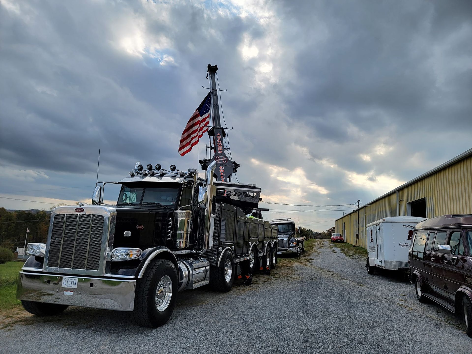 Black semi-truck with crane and American flag, parked on gravel path under cloudy sky.