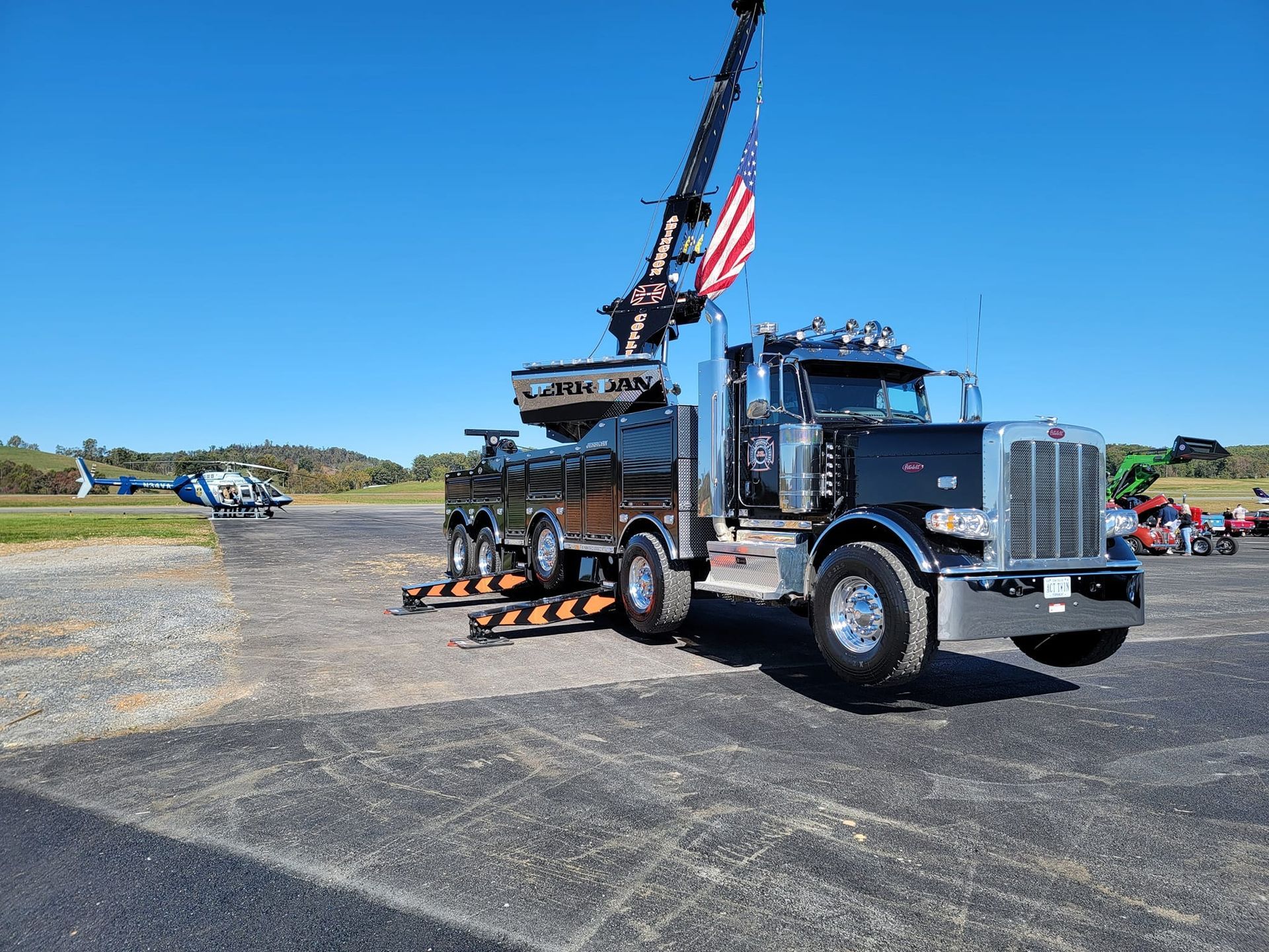 Black tow truck with crane, American flag, parked on asphalt. Helicopter and field in the background.