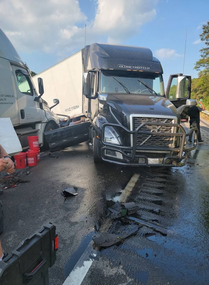 Two semi-trucks involved in a collision on a wet road. A person stands near the damaged trucks, scene likely outdoors.