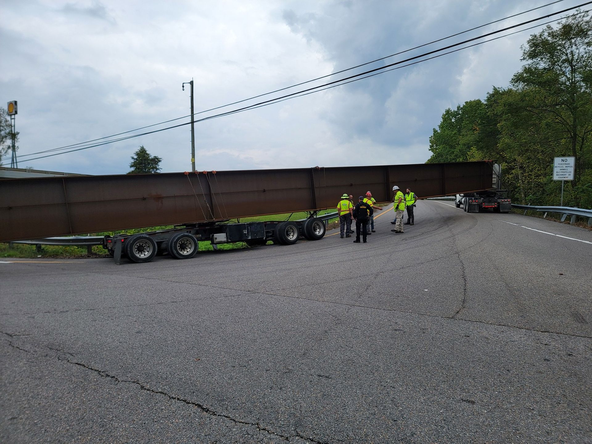 Semi-truck trailer blocking road; emergency responders at scene. Cloudy sky, asphalt road.