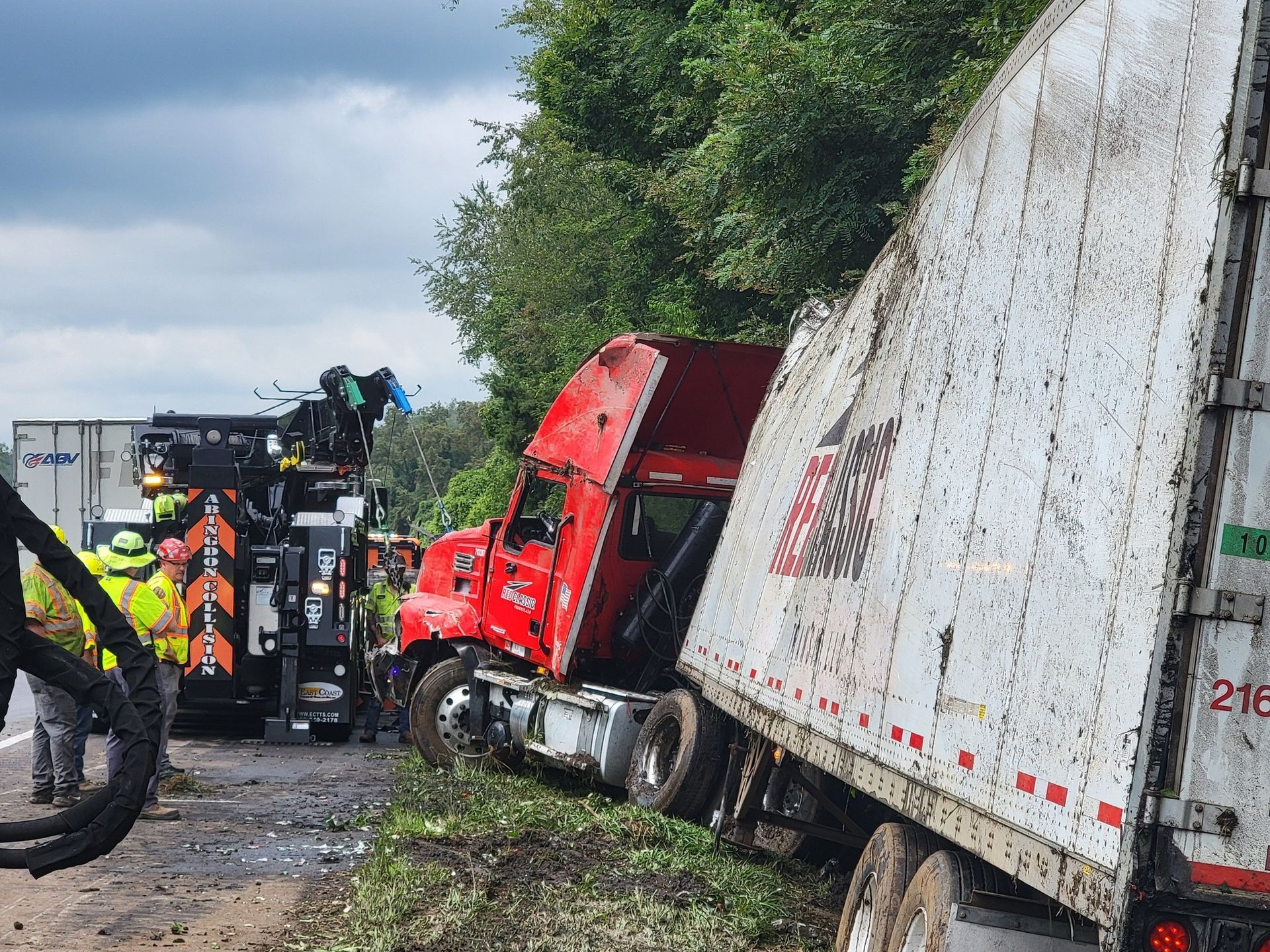 Red semi-truck crashed off the road, trailer damaged. Tow truck present, workers in safety vests.