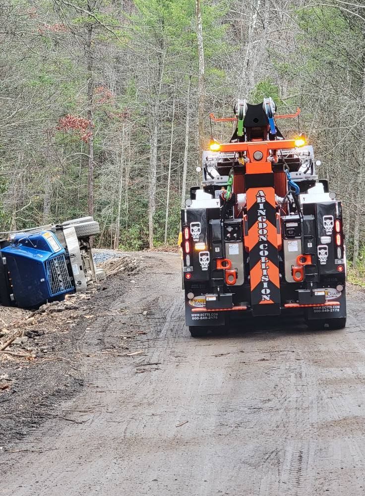 Tow truck facing a blue semi-truck overturned on a dirt road in a wooded area.