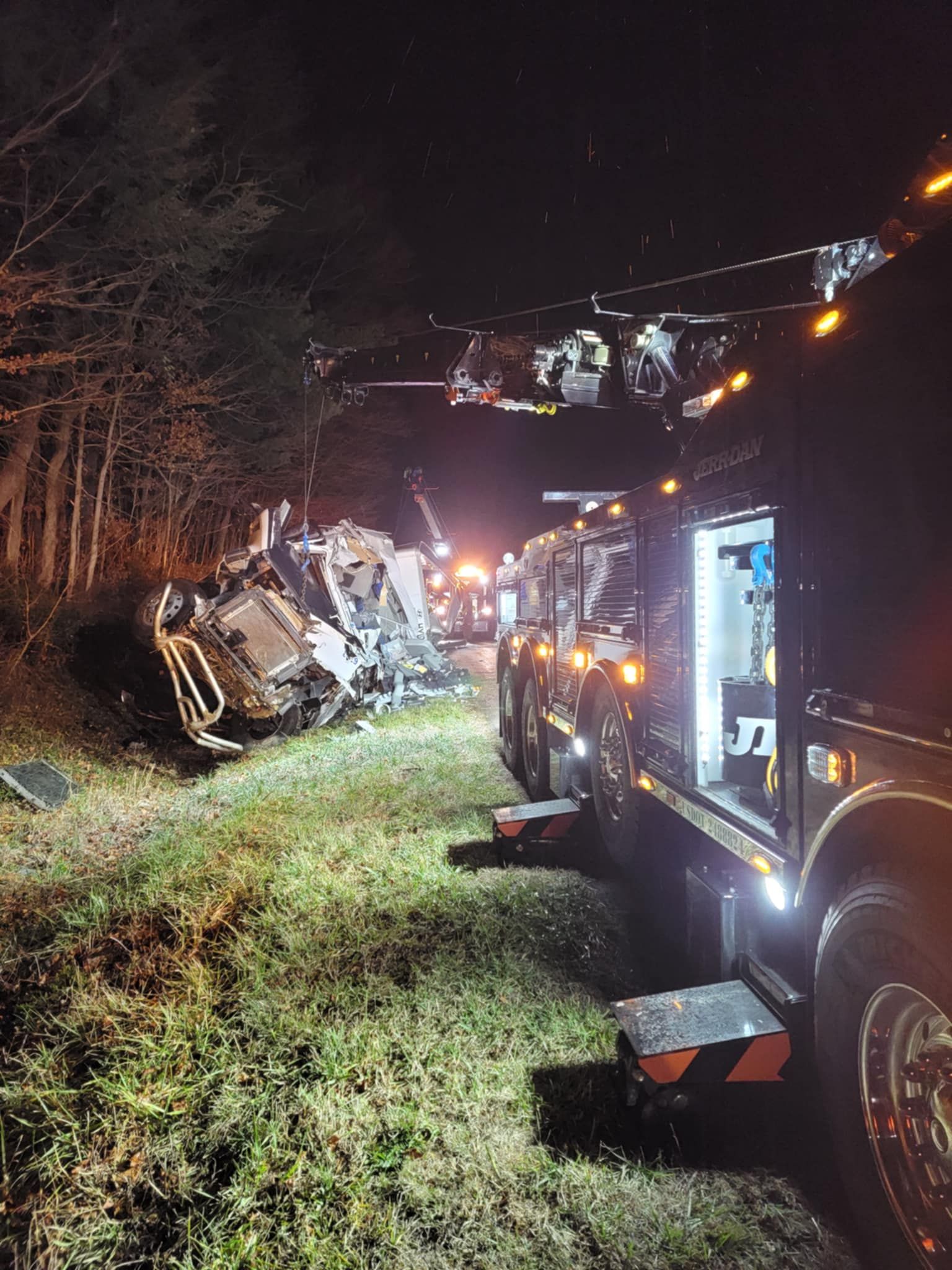 Tow trucks at night scene, extracting a severely damaged vehicle from ditch beside a wooded area.