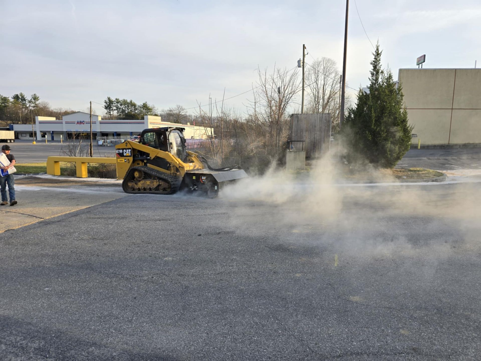 Yellow skid steer churning up dust on asphalt surface. Two people watch in the background.