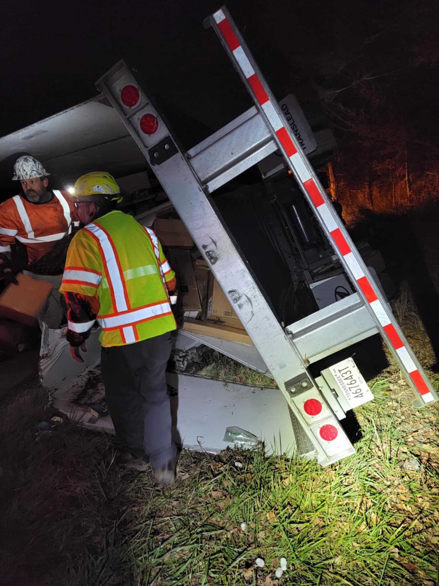 Two workers inspect a damaged silver trailer on grass, at night. One wears a reflective vest.