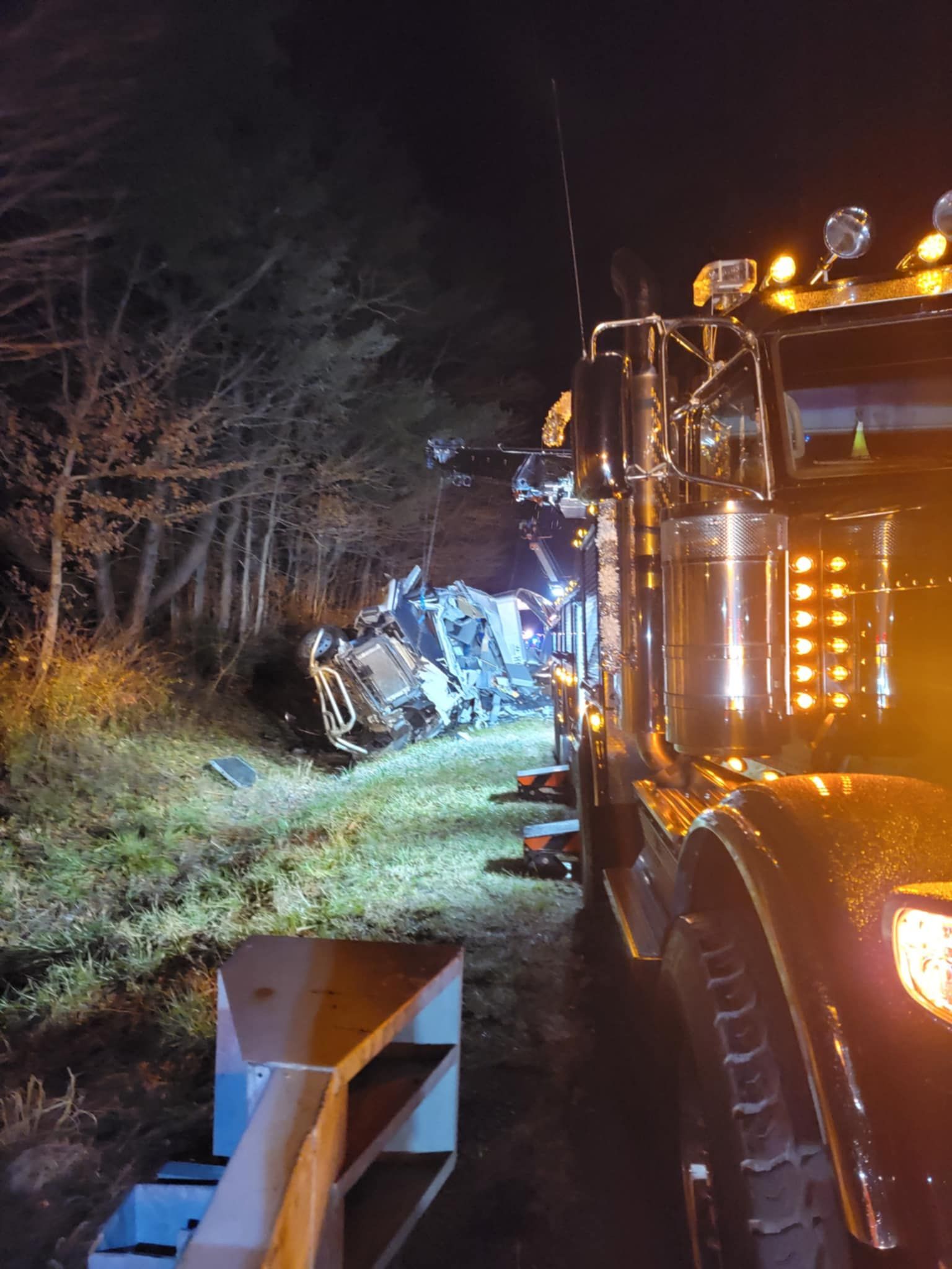Semi-truck parked at night, wreckage visible off-road. Trees line the left. Hazard lights illuminate.