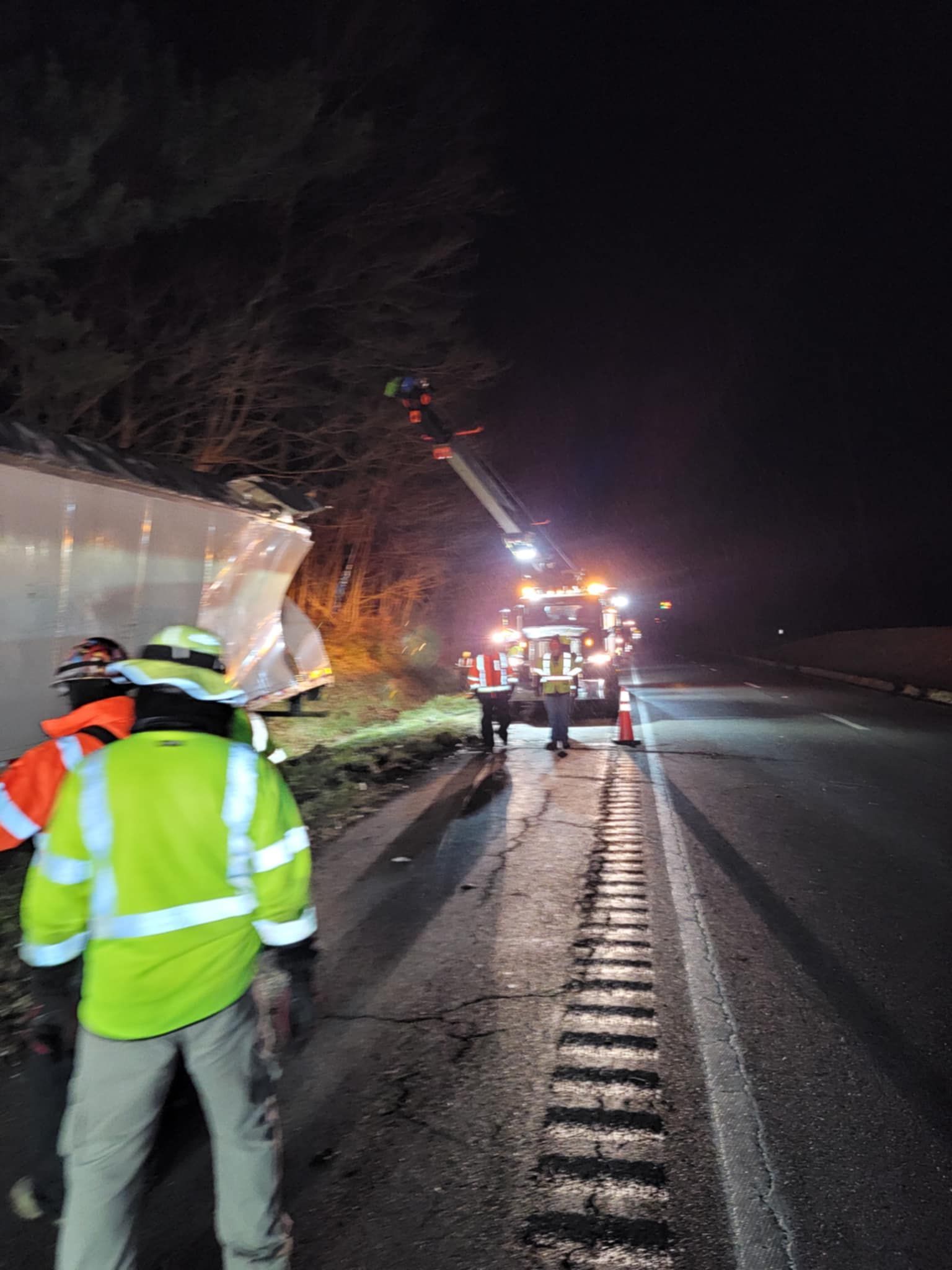 Night scene: tow truck with lights on, workers in vests near damaged barrier on roadside.