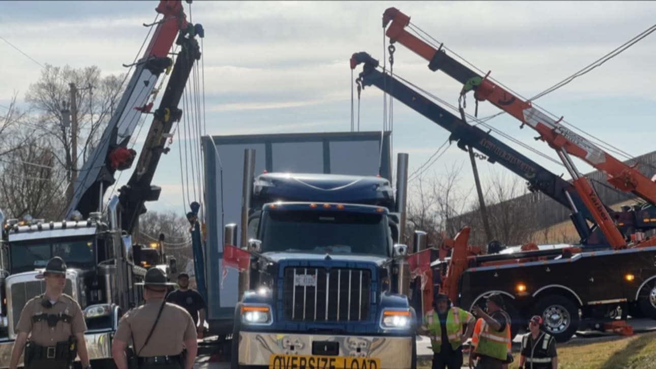 Semi-truck with an oversized load being assisted by two tow trucks; roadside.