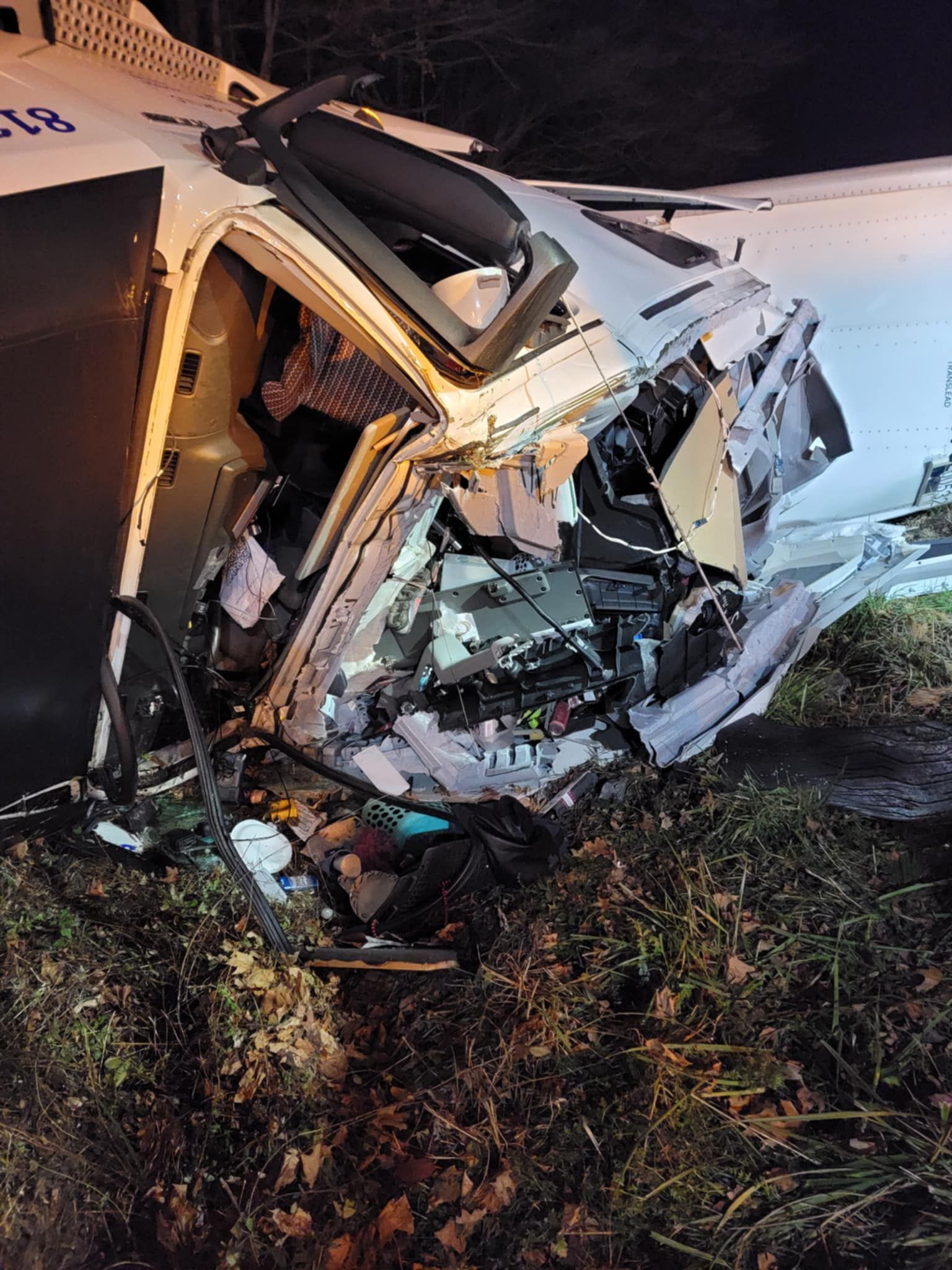 Severely damaged white vehicle on the side of a grassy area, front end crushed, night setting.