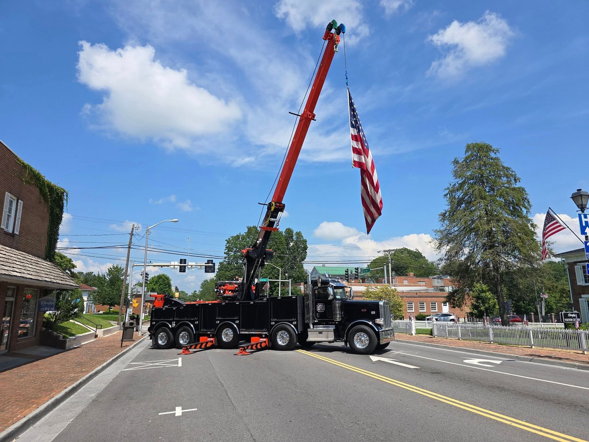 A crane truck raising an American flag on a street in front of buildings under a blue sky.