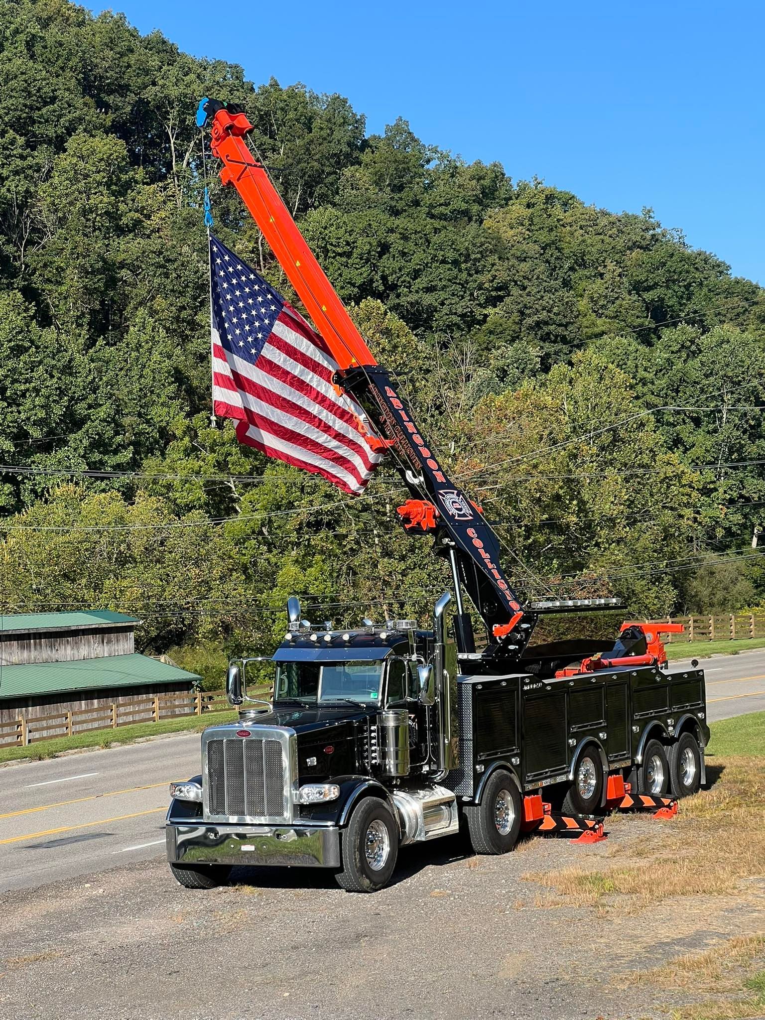 Black tow truck with extended orange boom, American flag, parked on roadside, trees in background.