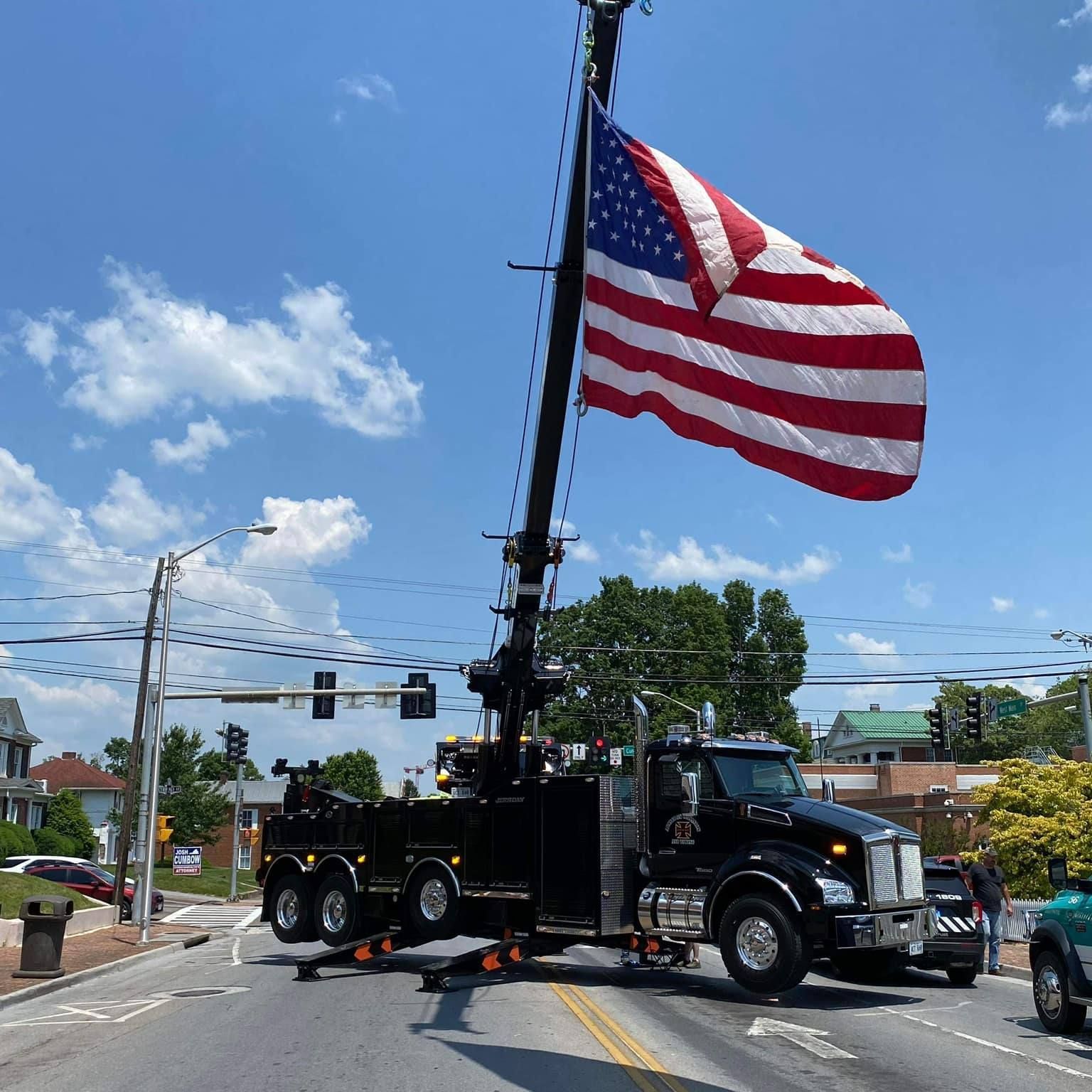 Black tow truck with US flag raised high in the air, on a city street with blue sky.