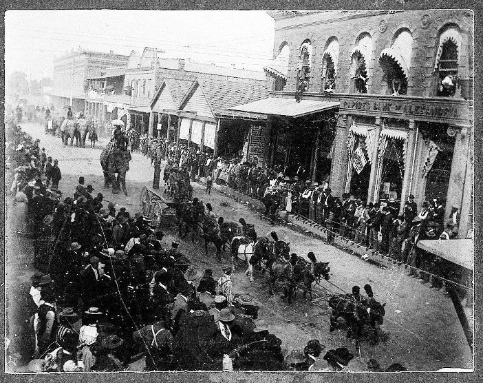A black and white photo of a parade with horse drawn carriages