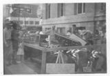A black and white photo of a group of people standing around a table in front of a building.
