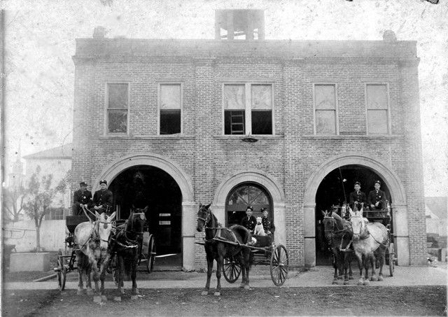 A black and white photo of a horse drawn carriage in front of a brick building.