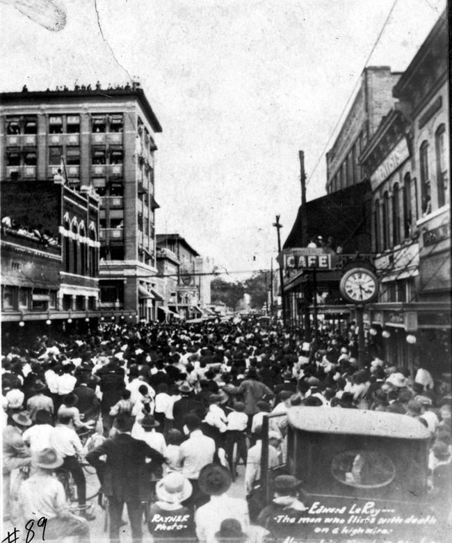 A black and white photo of a crowd of people in front of a cafe sign