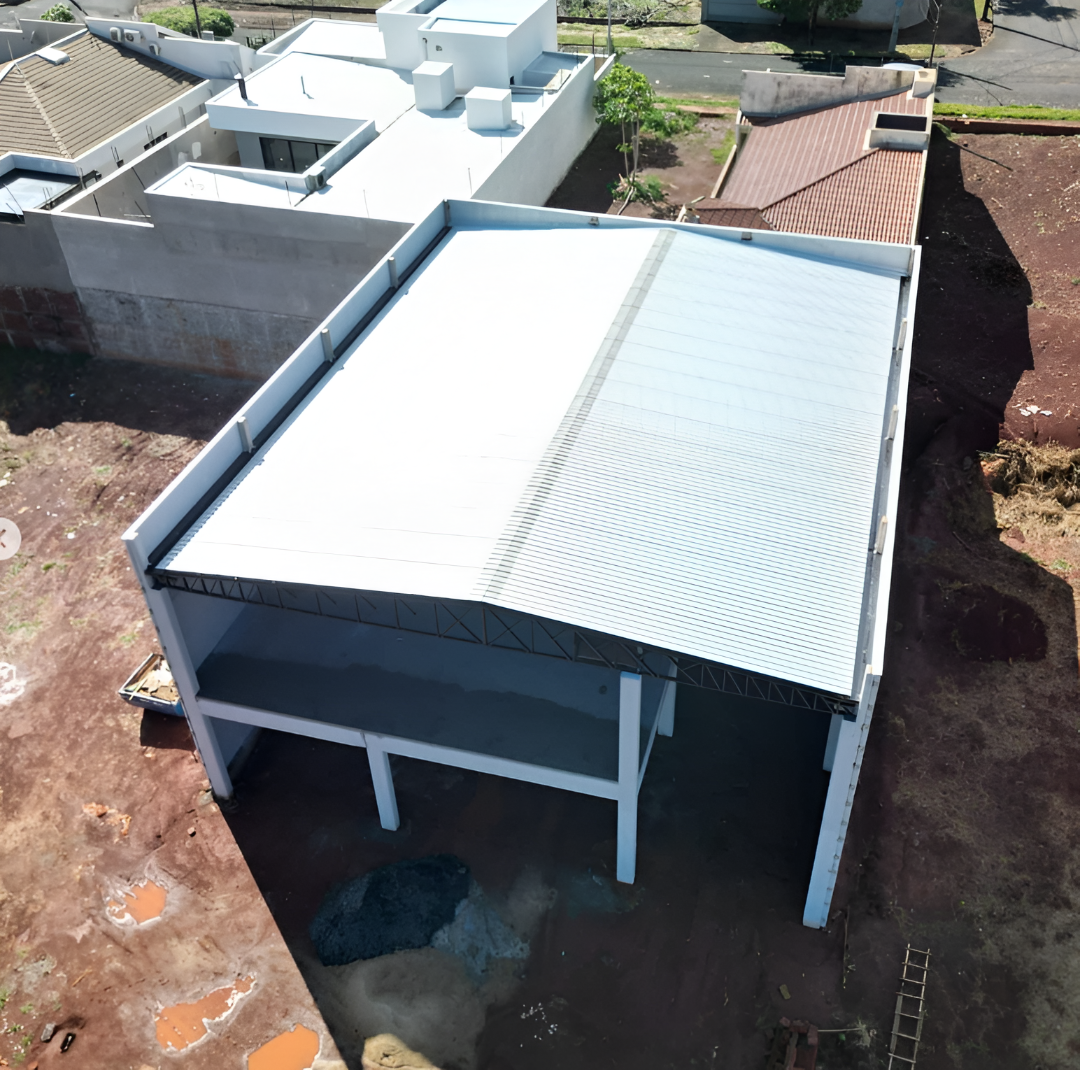 Vista aérea de uma obra, com telhado de metal, em construção num terreno de terra batida perto de casas residenciais.