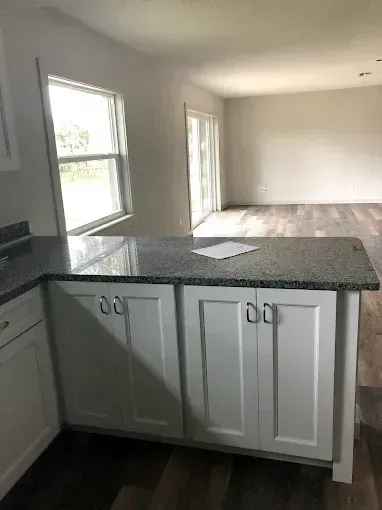 Empty open-concept kitchen and living area with white cabinets, granite counter, and two windows with daylight.