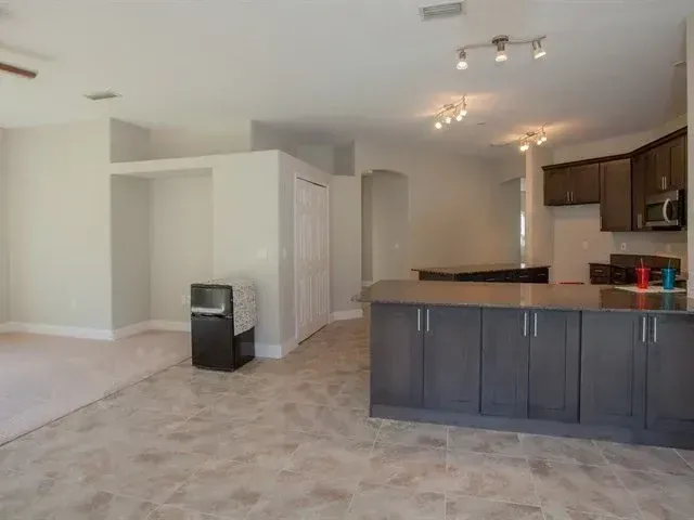 Empty open-plan kitchen and living area with dark cabinets, beige carpet, and tiled floor。