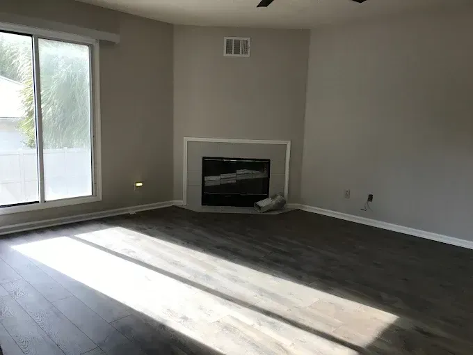 Empty living room with dark floors, gray walls, fireplace, and sunlight from sliding glass door.