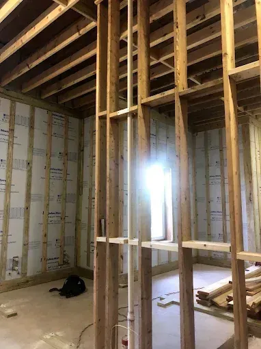Unfinished interior framing with exposed wooden studs and beams, sunlight through a window opening.