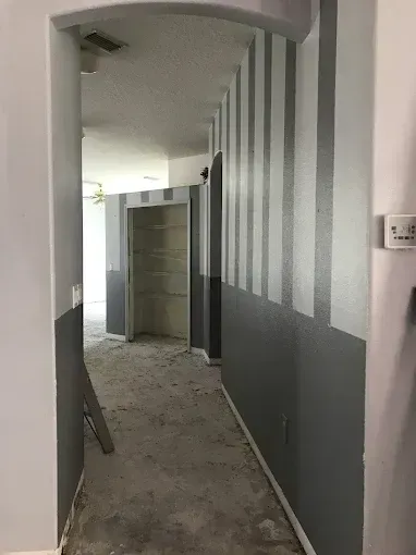 Narrow hallway under renovation with gray walls, debris on the floor, and an open door at the end