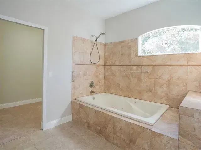 Empty beige-tiled bathroom with a built-in bathtub and tiled shower area, seen through an open doorway