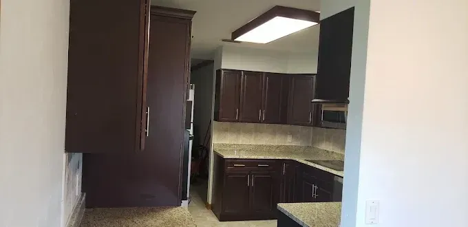 Small dark-wood kitchen with beige countertops, cabinets, and ceiling lights, viewed through a doorway.