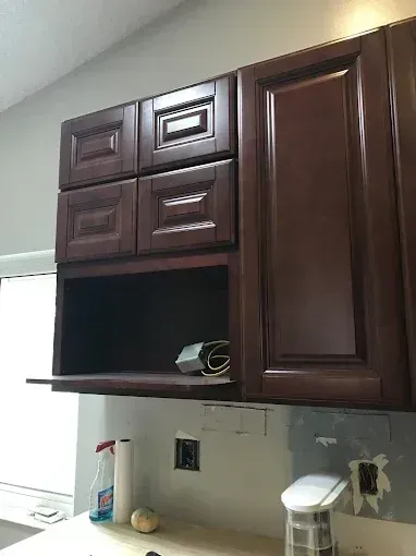 Dark wood kitchen cabinets above a countertop with cleaning supplies and an empty open shelf.