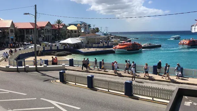 Busy seaside promenade with people, boats in a blue harbor, and sunny buildings along the waterfront
