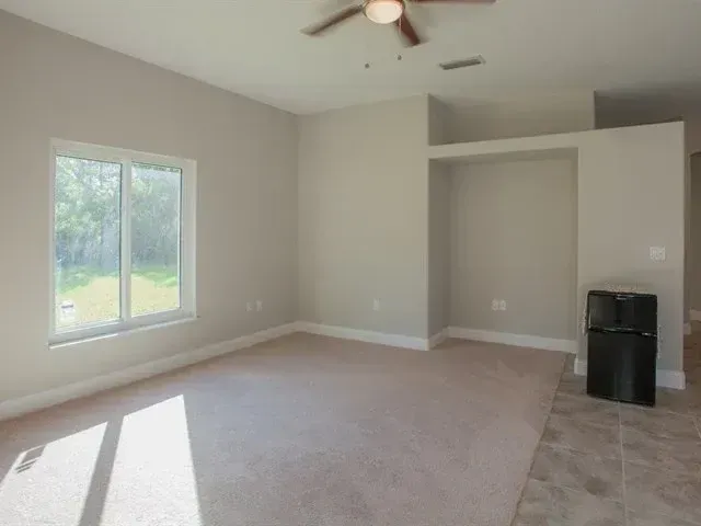 Empty carpeted living room with large window, ceiling fan, and tiled nook beside an open closet