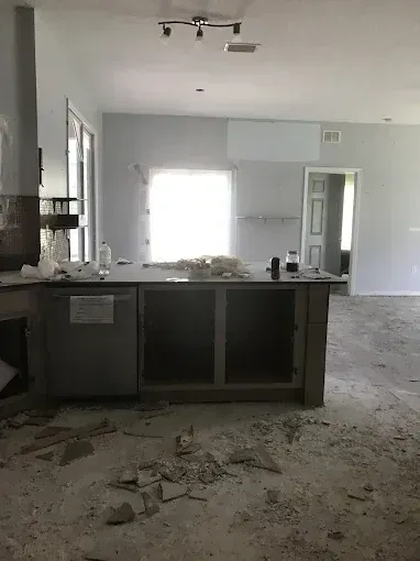 Renovated kitchen with dark island cabinets, debris on floor, and bright window light.